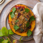 A halved vegan stuffed acorn squash filled with a quinoa stuffing, garnished with parsley and sage leaves, sits on a ceramic plate next to a white napkin and some green leaves.