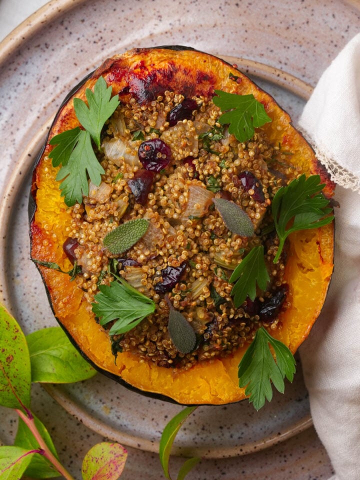 A halved vegan stuffed acorn squash filled with a quinoa stuffing, garnished with parsley and sage leaves, sits on a ceramic plate next to a white napkin and some green leaves.