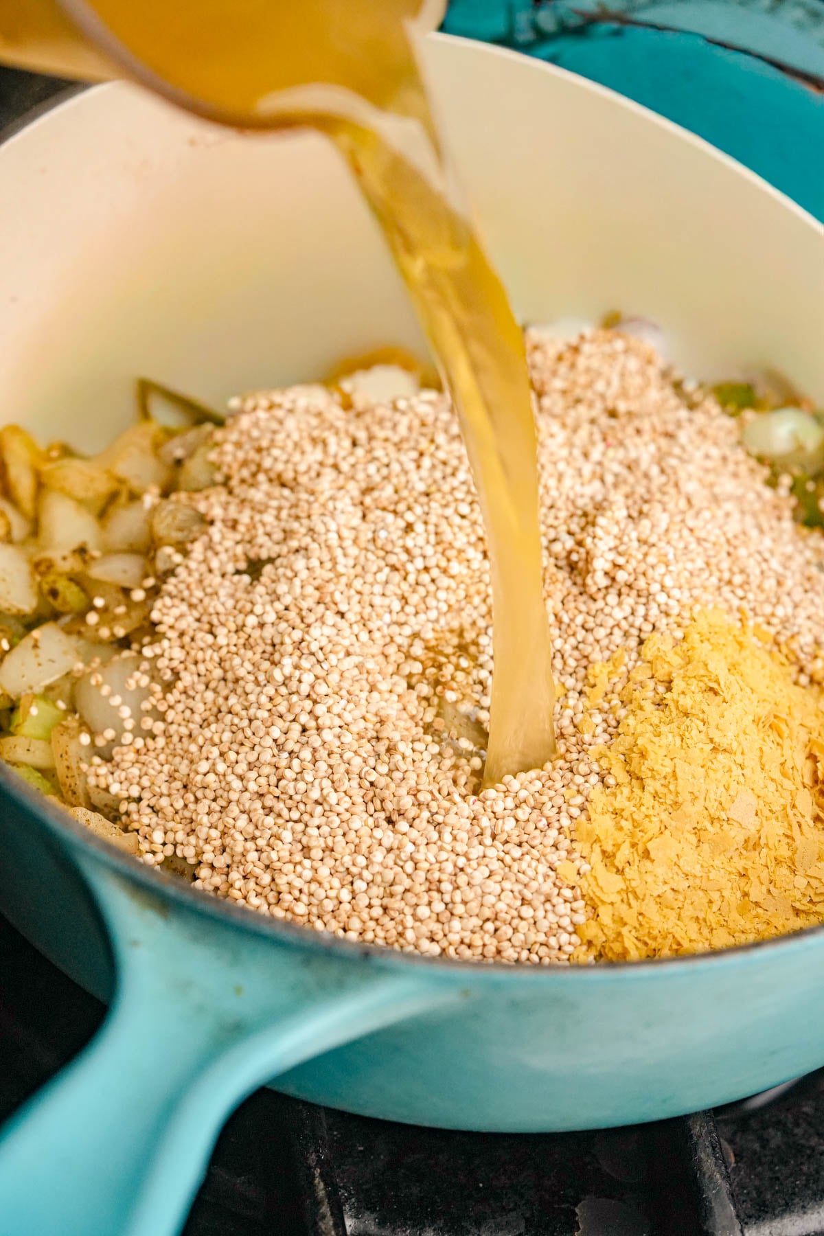Broth is being poured into a blue pot containing uncooked quinoa, chopped onions, celery, and a pile of yellow seasoning, likely nutritional yeast.