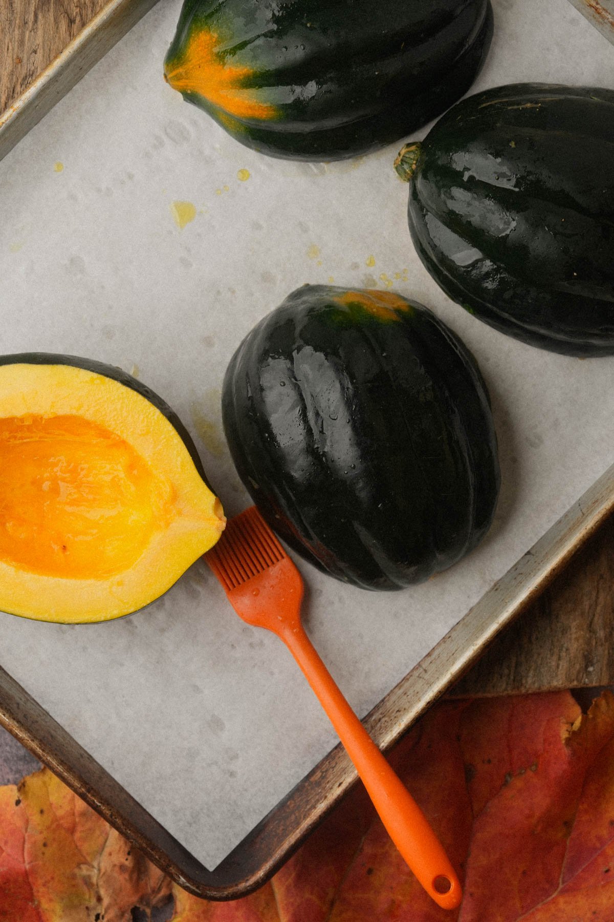 Three whole acorn squashes and one halved acorn squash on a parchment-lined baking sheet with an orange basting brush, set on a wooden surface with autumn leaves.
