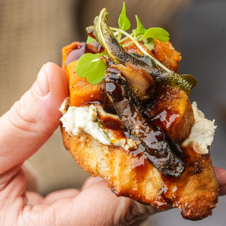 A hand holds a toasted butternut squash crostini with creamy cheese, roasted butternut squash, mushrooms, microgreens, crispy sage, and a drizzle of dark balsamic glaze.