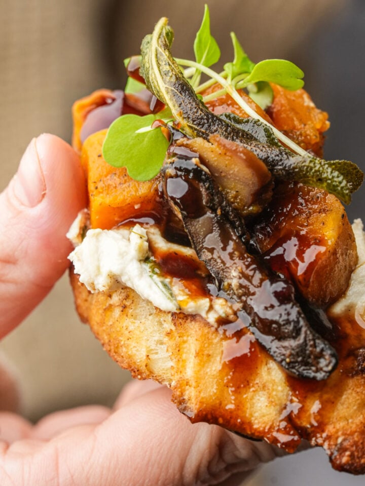 A hand holds a toasted butternut squash crostini with creamy cheese, roasted butternut squash, mushrooms, microgreens, crispy sage, and a drizzle of dark balsamic glaze.