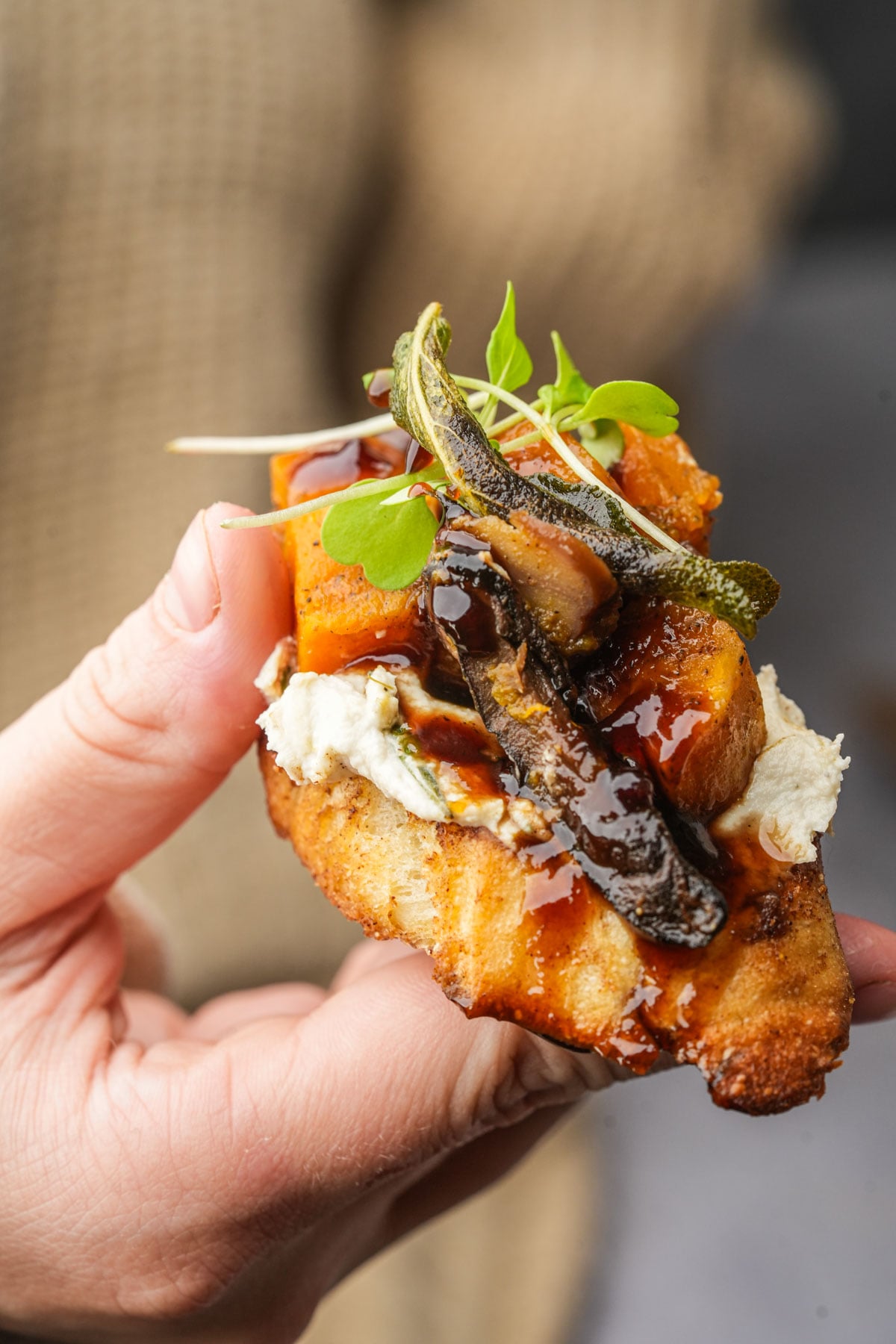 A hand holds a piece of butternut squash crostini topped with roasted vegetables, creamy cheese, microgreens, and a drizzle of dark sauce, against a blurred background.