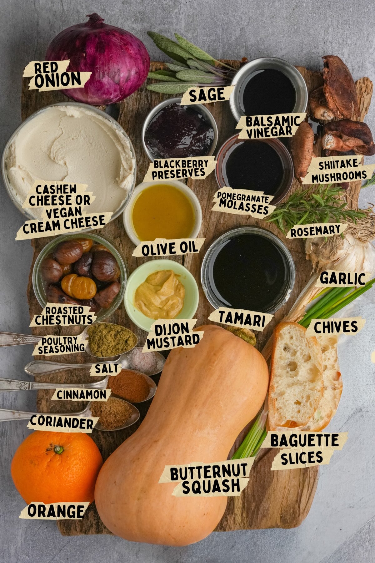 A wooden board displays labeled ingredients for a butternut squash crostini recipe including butternut squash, orange, red onion, cashew cheese, olive oil, seasonings, herbs, mustard, roasted chestnuts, mushrooms, and baguette slices.