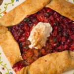 A rustic cranberry galette with a golden-brown crust, filled with baked cranberries and topped with a dollop of whipped cream sprinkled with cinnamon, served on a decorated plate.