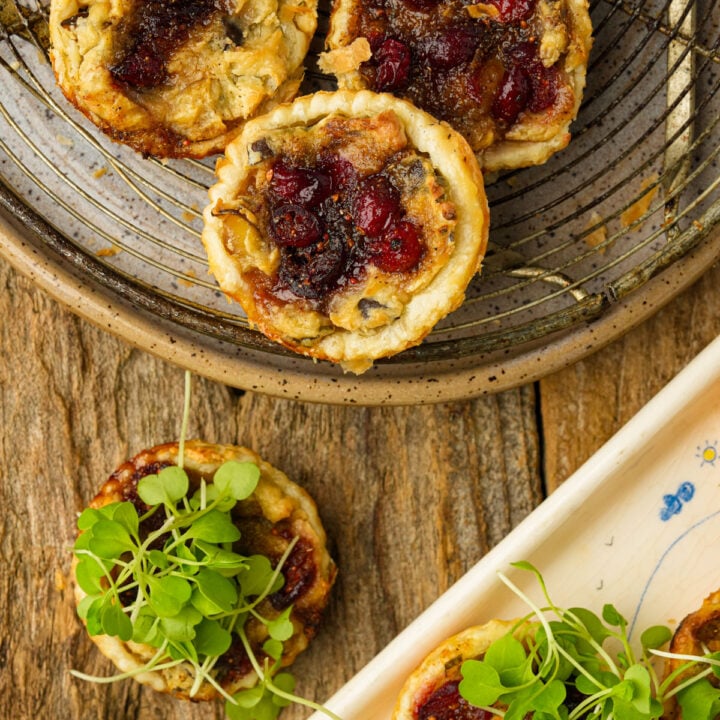 Small savory cranberry puff pastries, some topped with cranberries garnished with fresh green microgreens, displayed on rustic ceramic and wooden plates on a wooden surface.