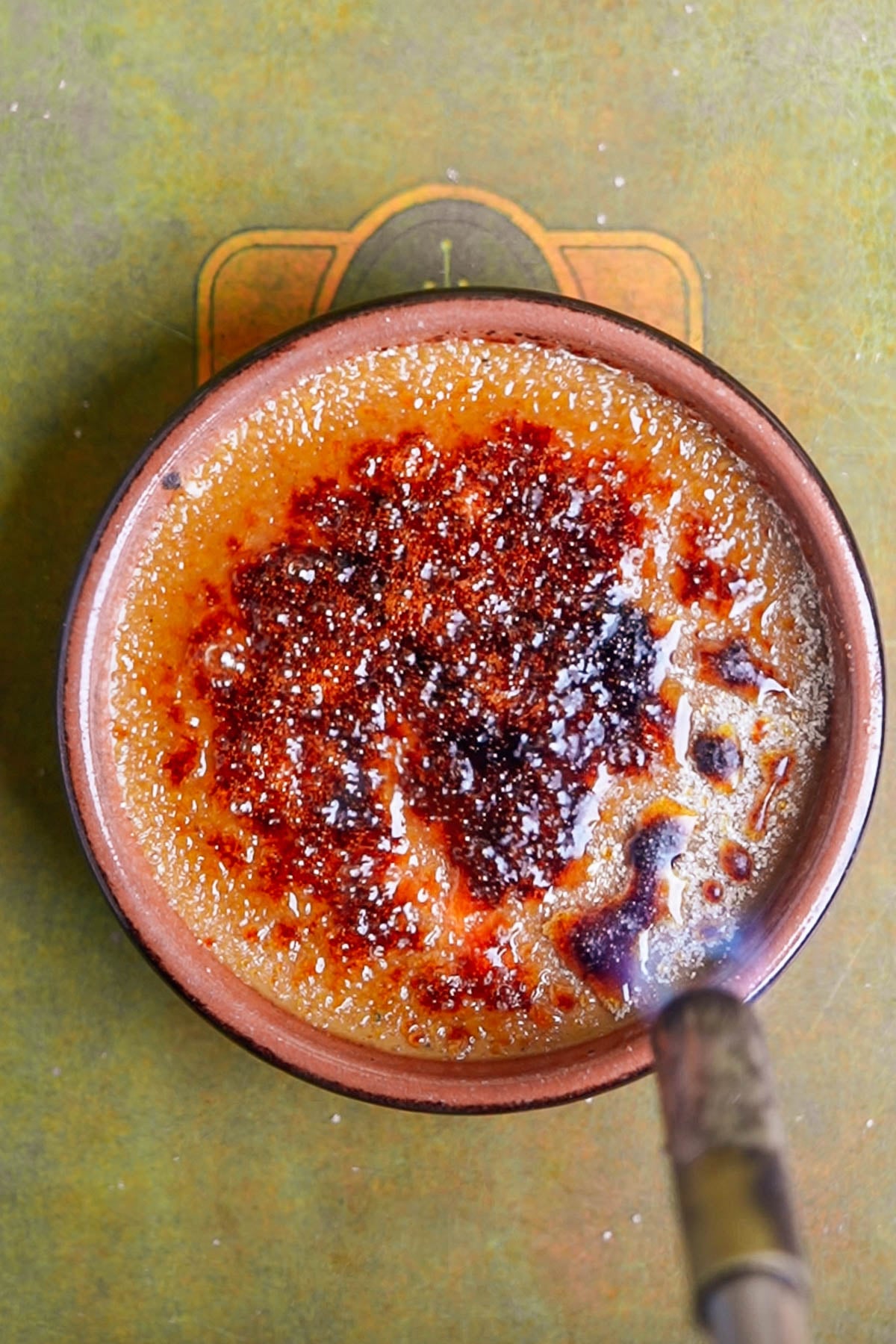 A close-up of a pumpkin creme brulee in a brown dish, with a caramelized sugar crust being torched, creating a golden and dark brown surface on top of the creamy dessert.