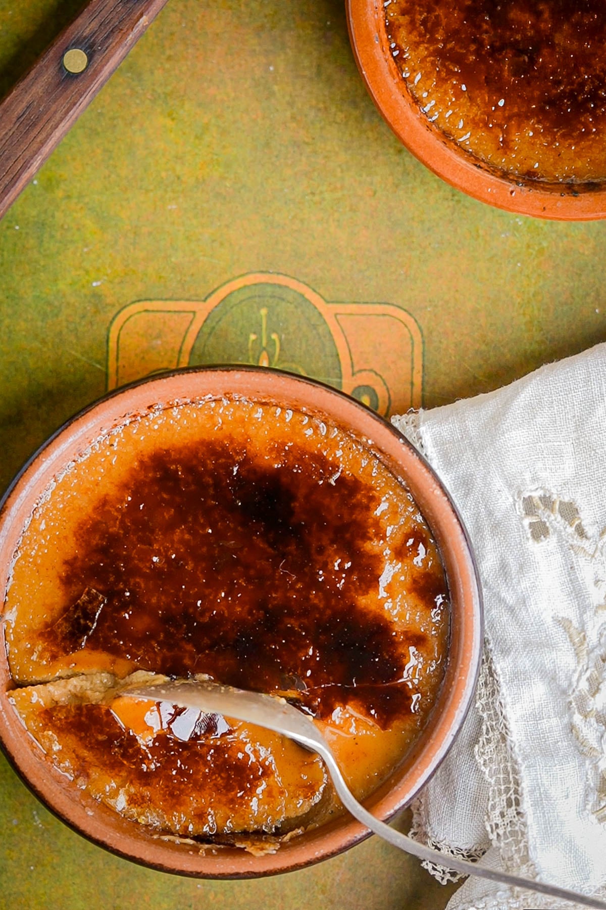 A close-up of two pumpkin creme brulee desserts in brown ramekins, one with a spoon breaking the caramelized sugar crust. A vintage lace napkin lies nearby on a textured green surface.