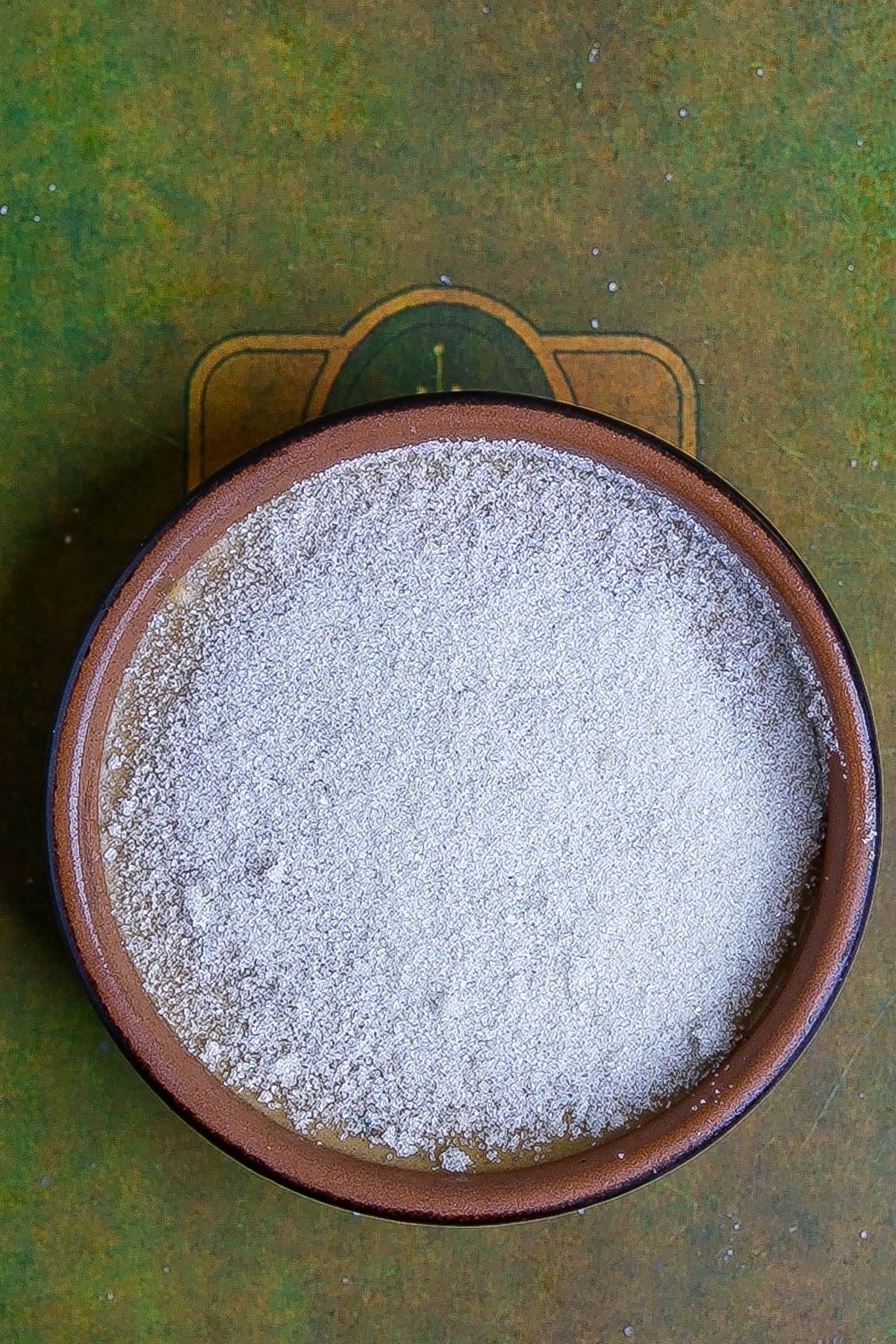 A brown ceramic bowl filled with fine white powder, possibly sugar for pumpkin creme brulee, sits on a greenish surface with a faint circular design underneath.