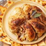 A golden-brown pumpkin puff pastry, shaped like a pumpkin and topped with a cinnamon stick stem and sage leaves, is served on a plate surrounded by triangular crackers.