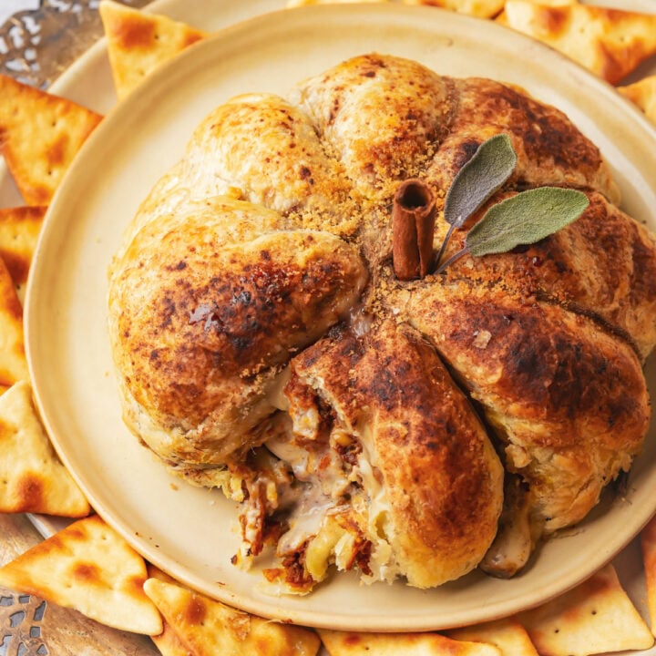 A golden-brown pumpkin puff pastry, shaped like a pumpkin and topped with a cinnamon stick stem and sage leaves, is served on a plate surrounded by triangular crackers.