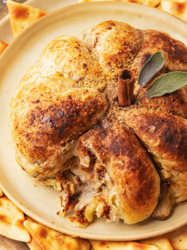 A golden-brown pumpkin puff pastry, shaped like a pumpkin and topped with a cinnamon stick stem and sage leaves, is served on a plate surrounded by triangular crackers.