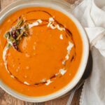 A bowl of creamy Thai pumpkin curry soup garnished with a swirl of coconut milk a drizzle of kecap manis, and topped with fried chilies and herbs, placed on a wooden surface next to a white cloth napkin and a metal spoon.