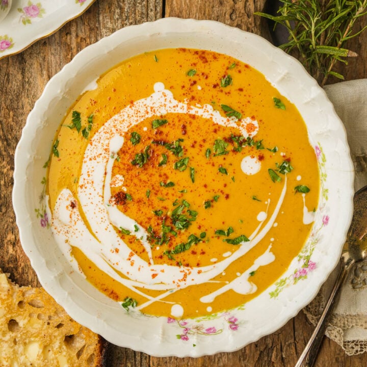 A bowl of creamy orange vegan butternut squash soup garnished with a swirl of coconut milk, chopped herbs, and a sprinkle of red spices, set on a rustic wooden table with a slice of bread and fresh herbs nearby.