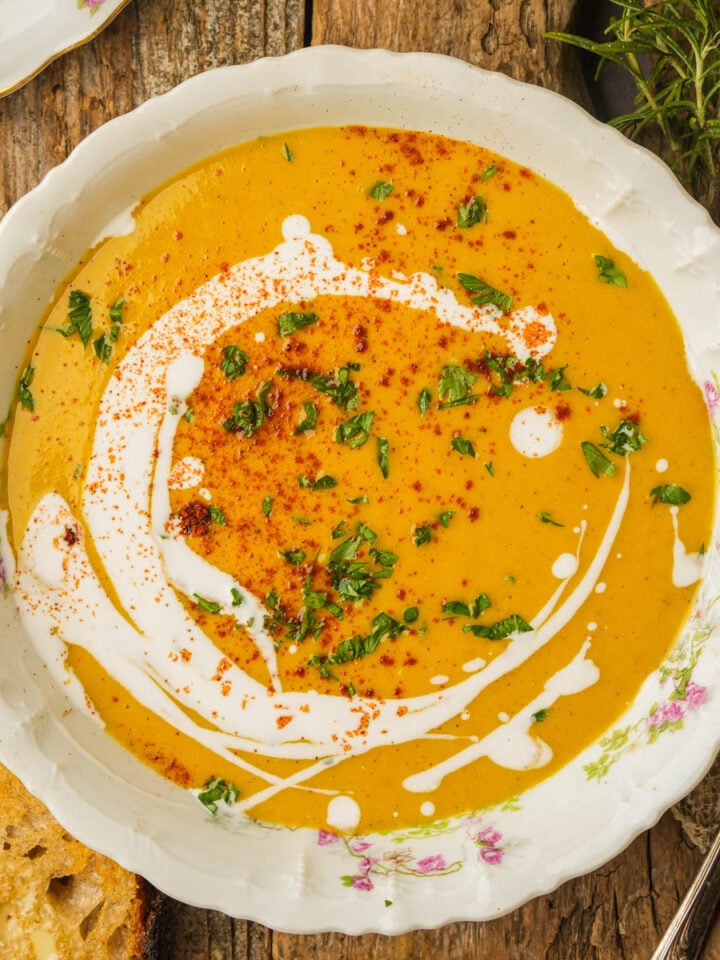 A bowl of creamy orange vegan butternut squash soup garnished with a swirl of coconut milk, chopped herbs, and a sprinkle of red spices, set on a rustic wooden table with a slice of bread and fresh herbs nearby.