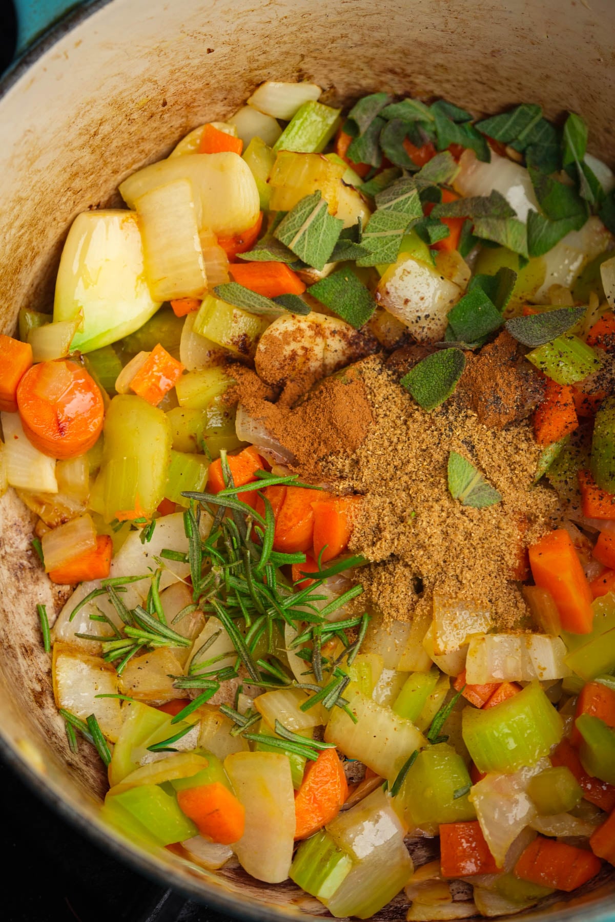 Chopped onions, carrots, and celery cooking in a pot with fresh rosemary and sage leaves, plus ground spices piled in the center.