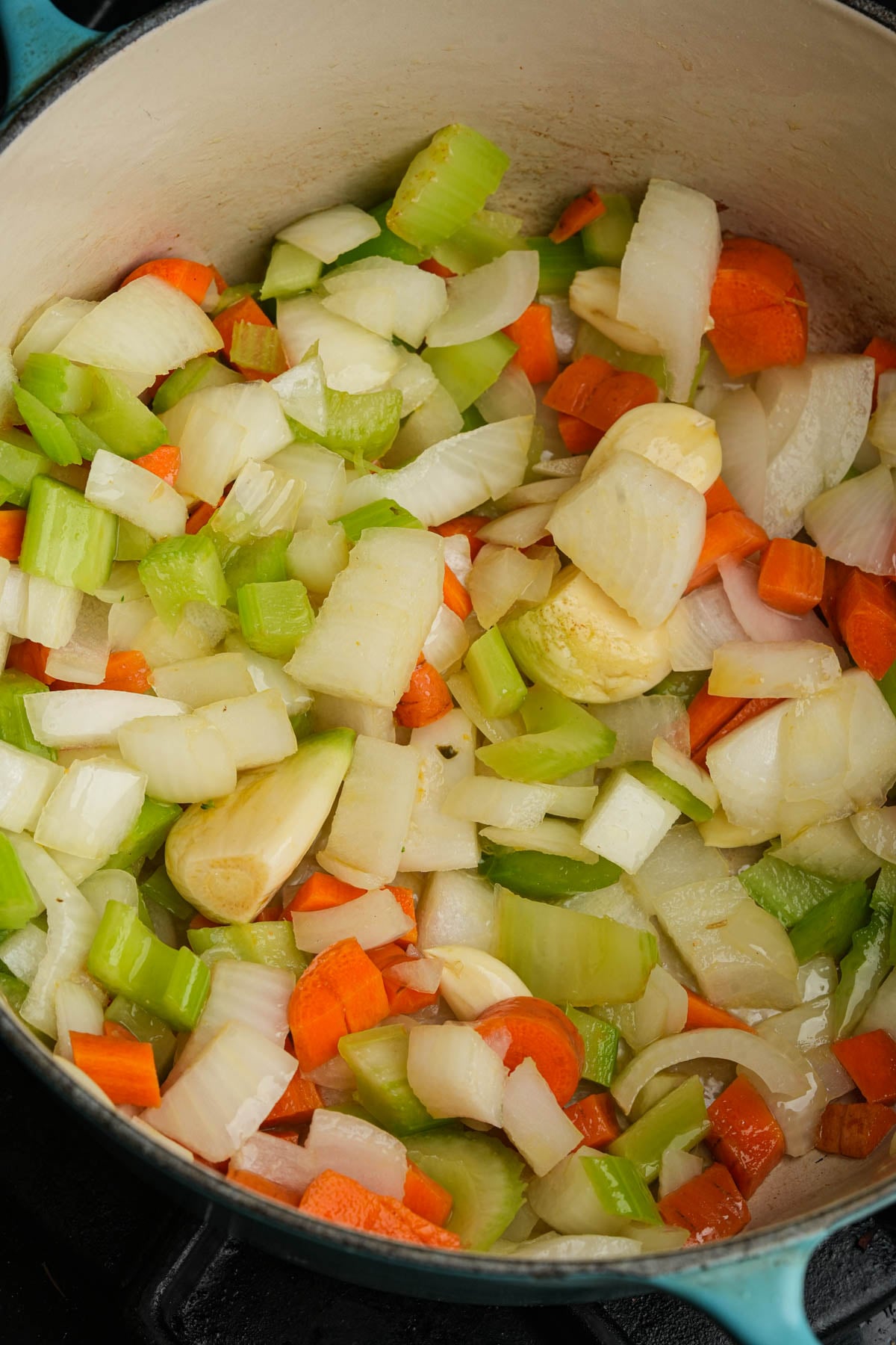 Chopped onions, carrots, celery, and whole garlic cloves cooking in a pot, forming the base for a vegan butternut squash soup.