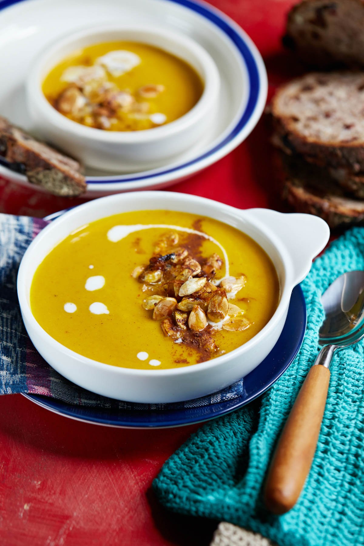 Two bowls of creamy vegan butternut squash soup topped with seeds and a swirl of cream, surrounded by slices of rustic bread on a red table, next to a spoon and a textured teal napkin.