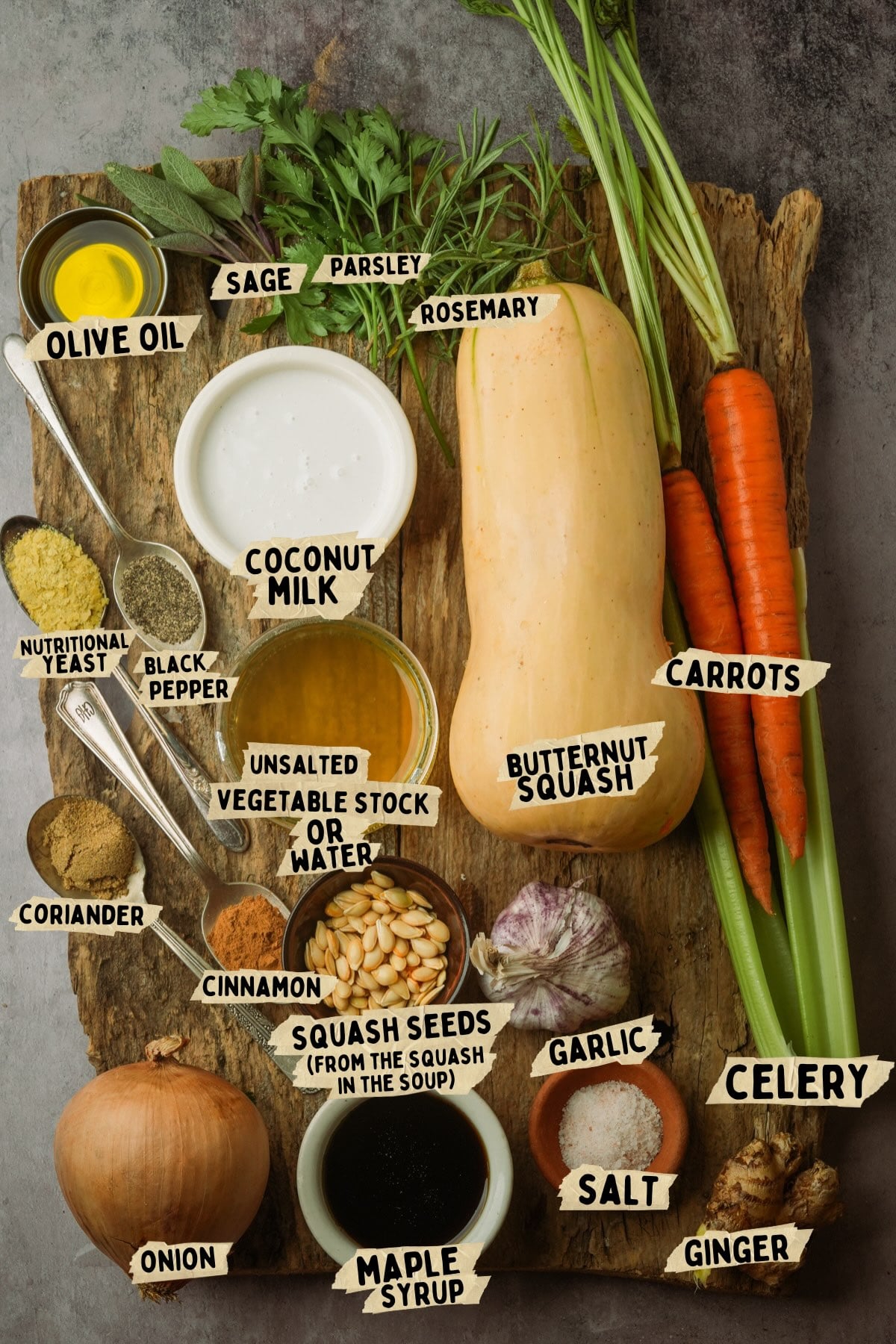 A wooden board displaying ingredients for a vegan butternut squash soup recipe including butternut squash, carrots, celery, onion, garlic, ginger, olive oil, coconut milk, stock, nutritional yeast, spices, salt, maple syrup, and fresh herbs.