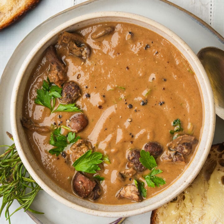 A bowl of creamy brown roasted mushroom soup with chunks of mushroom garnished with fresh parsley and cracked black pepper, served with a slice of bread on the side.