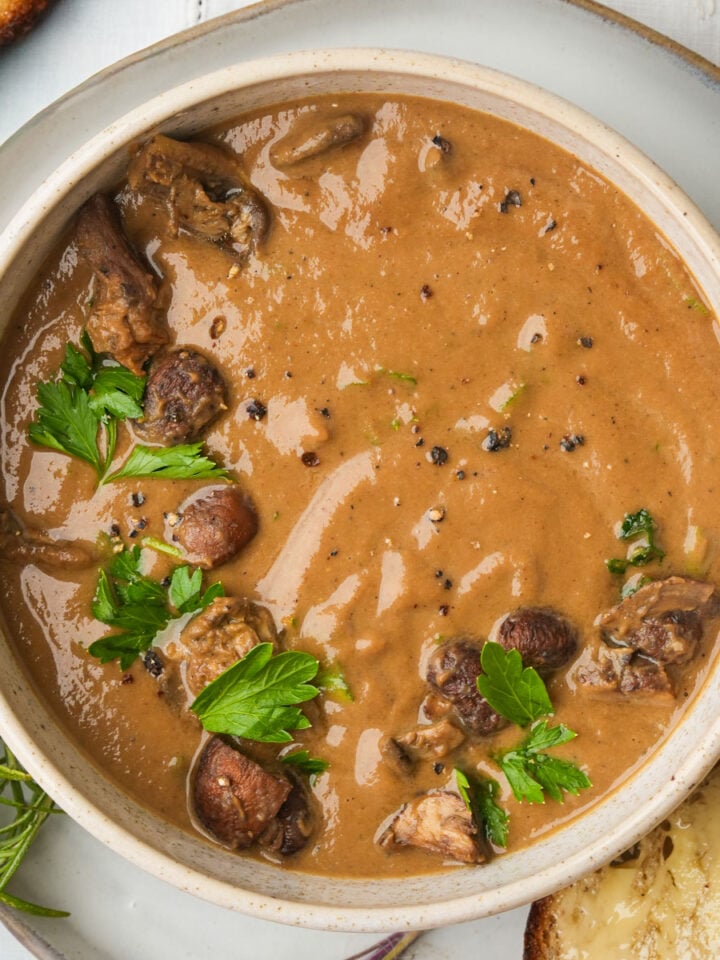 A bowl of creamy brown roasted mushroom soup with chunks of mushroom garnished with fresh parsley and cracked black pepper, served with a slice of bread on the side.