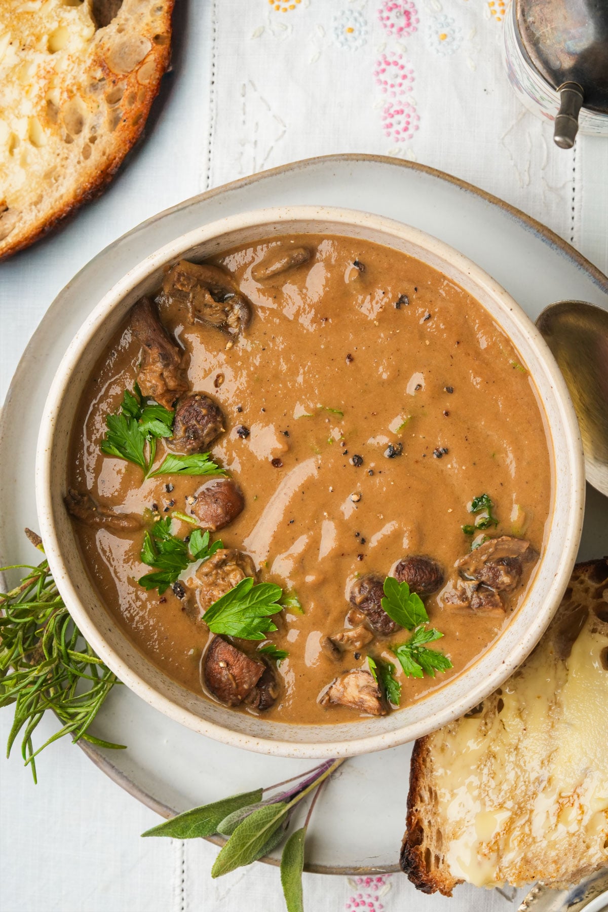 A bowl of creamy vegan mushroom soup garnished with fresh parsley and cracked pepper, served with slices of crusty buttered bread on a white plate, with herbs on the side.