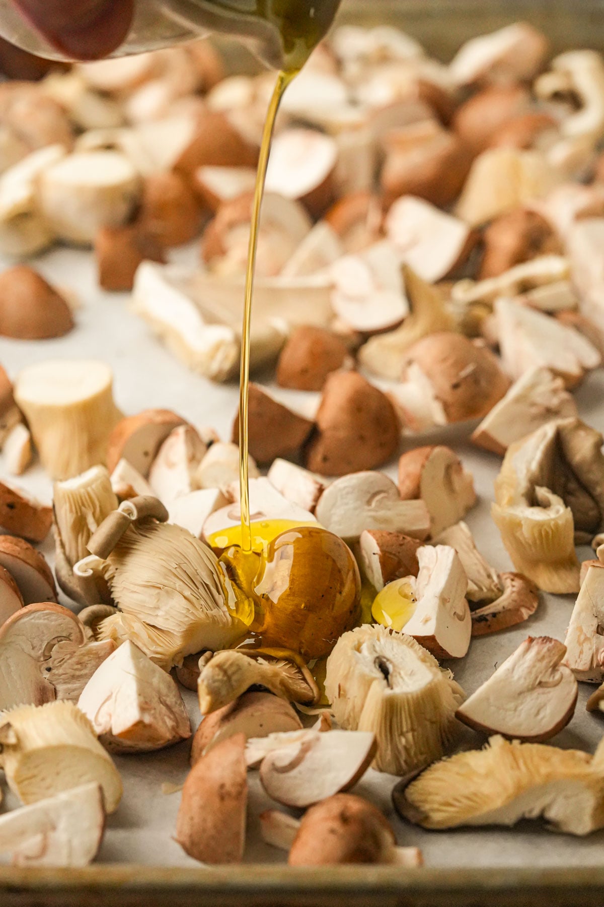 Chopped mushrooms spread on a baking sheet, with golden olive oil being poured over them from above.