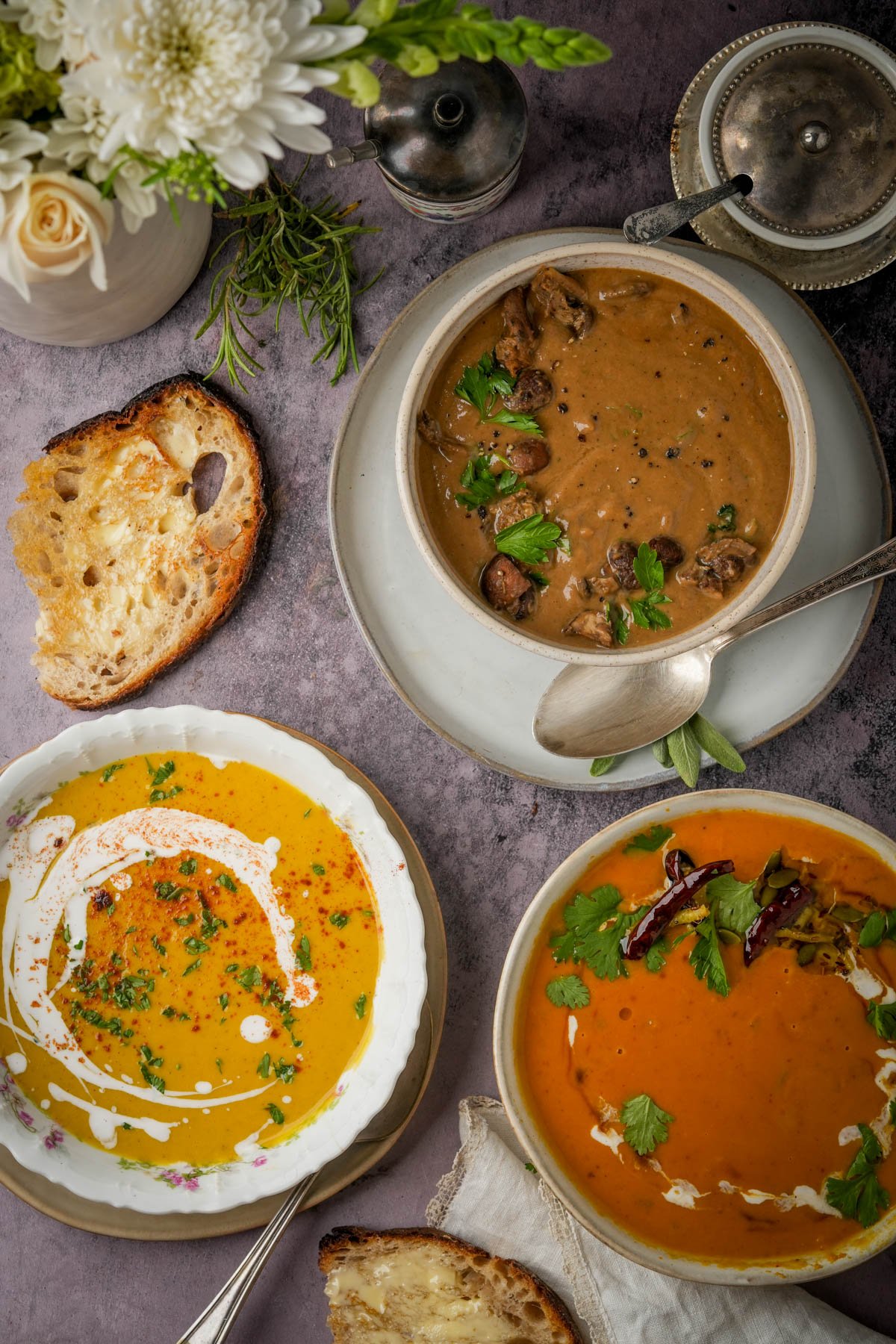 Three bowls of soup: roasted mushroom soup, vegan butternut squash soup, and Thai pumpkin curry soup are arranged on a purple surface with slices of toasted bread, fresh herbs, and a bouquet of white flowers nearby.