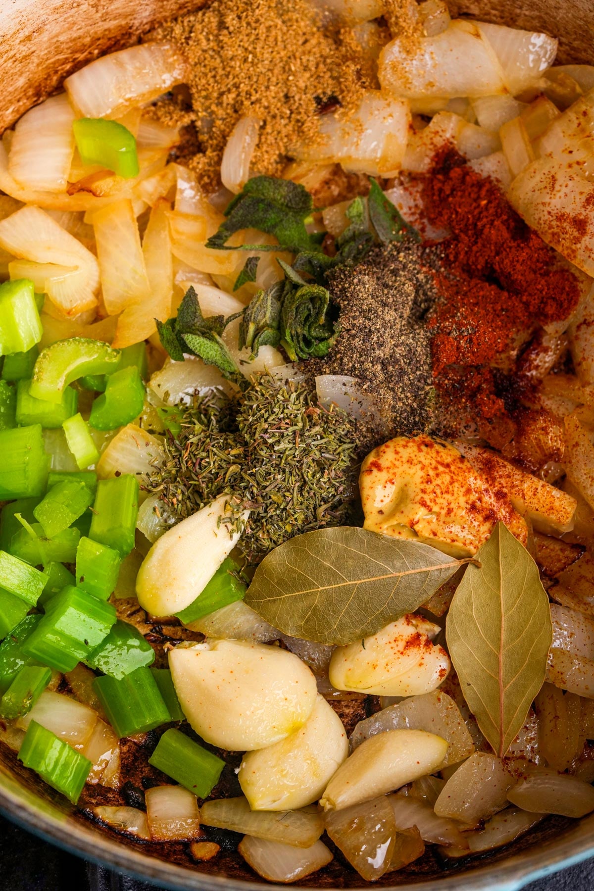 Chopped onions and celery, whole garlic cloves, bay leaves, and various dried herbs and spices are arranged in a pot, ready to be cooked.