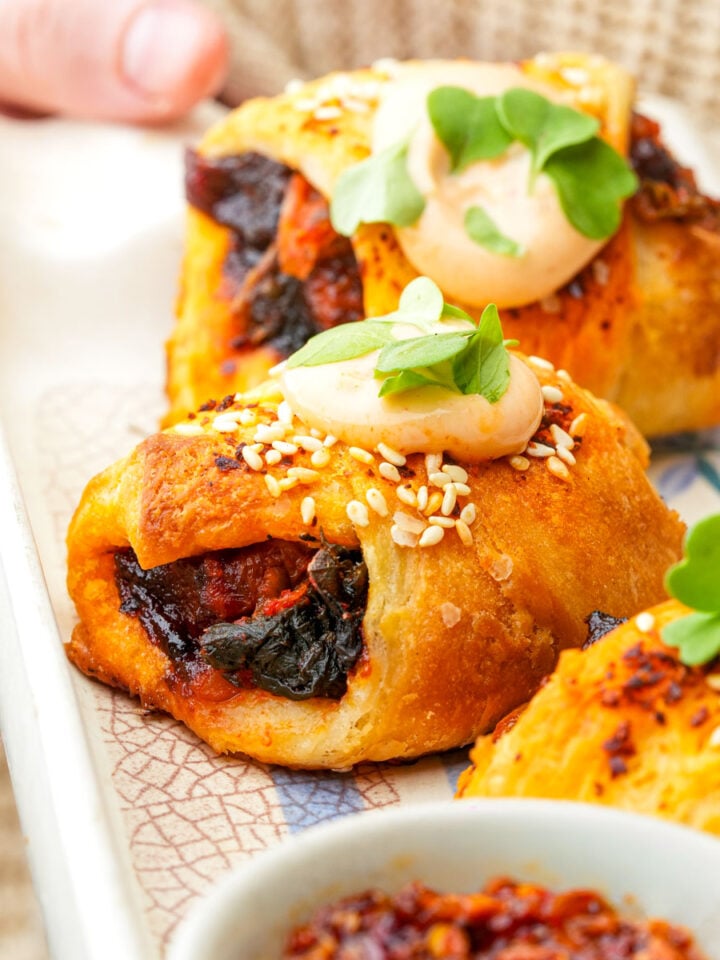 Golden, flaky vegan pigs in a blanket filled with smoked carrots and kimchi, topped with sesame seeds, creamy sauce, and fresh microgreens on a decorative plate.