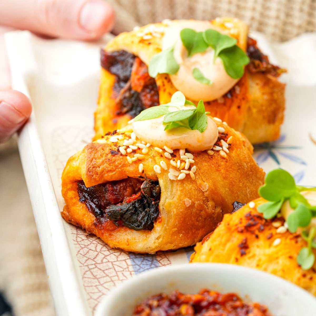 vegan_pigs_in_a_blanket-03 Golden, flaky vegan pigs in a blanket filled with smoked carrots and kimchi, topped with sesame seeds, creamy sauce, and fresh microgreens on a decorative plate.