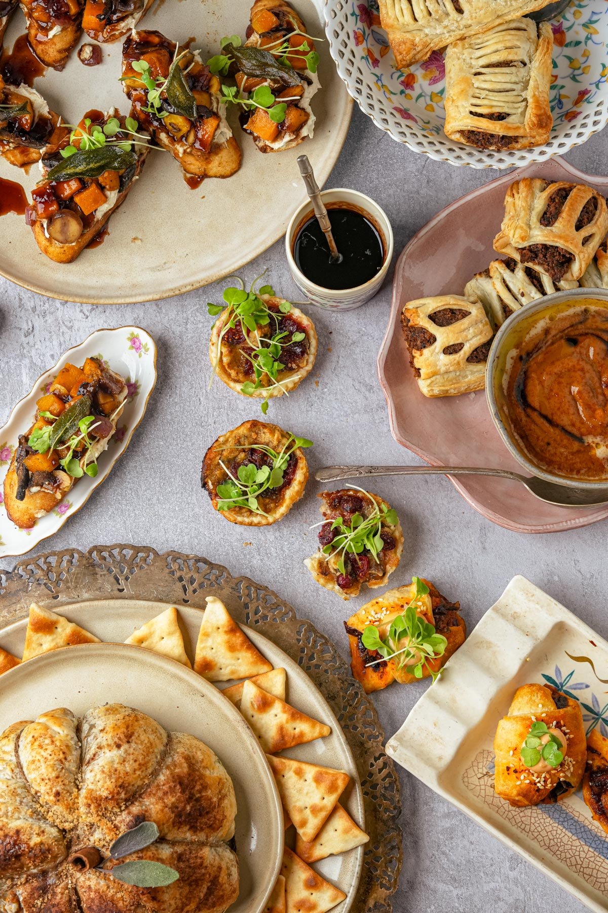 A table with assorted appetizers, including butternut squash crostini with toppings, vegan pigs in a blanket, pumpkin puff pastry, cranberry puff pastries, and vegan sausage rolls, all artfully arranged on various plates and trays.