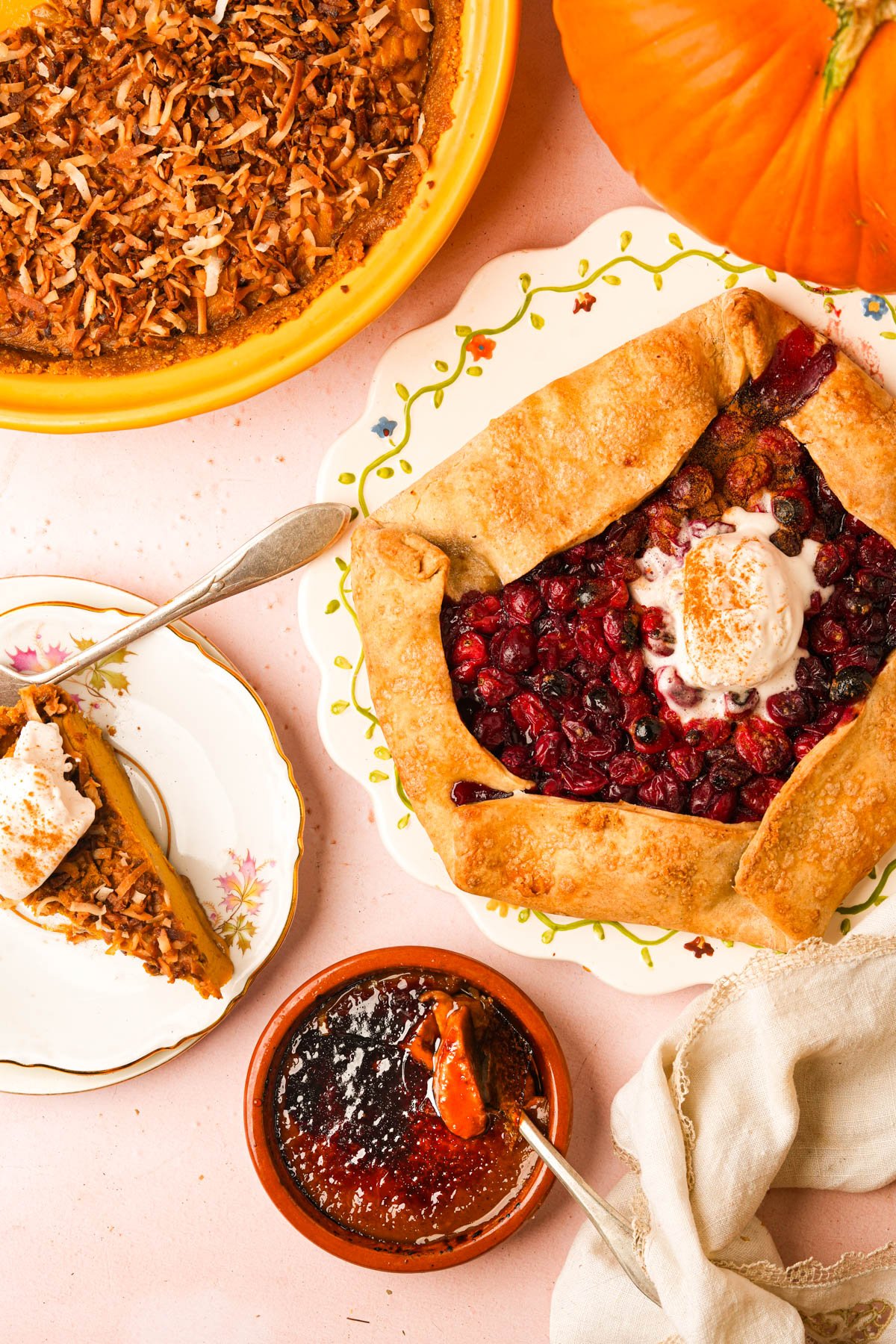 A rustic cranberry galette with ice cream, a slice of vegan pumpkin chiffon pie, a whole coconut pie, a serving of vegan pumpkin creme brulee, and a pumpkin are arranged on a pink tabletop, with serving utensils and a napkin nearby.