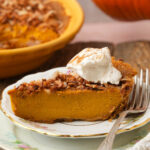 A slice of vegan pumpkin chiffon pie topped with toasted coconut and a dollop of whipped cream sits on a decorative plate with a fork. The rest of the pie is in the background.