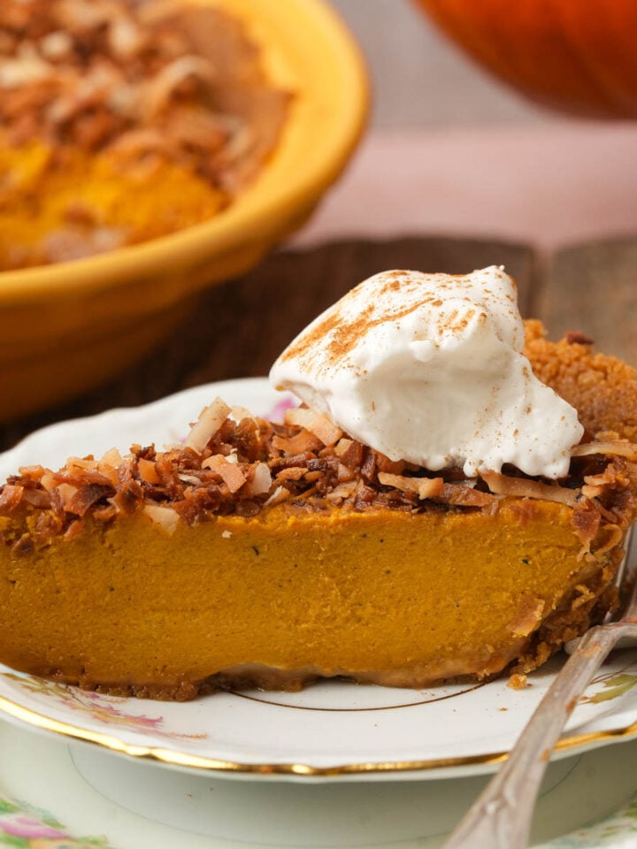 A slice of vegan pumpkin chiffon pie topped with toasted coconut and a dollop of whipped cream sits on a decorative plate with a fork. The rest of the pie is in the background.