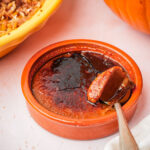 A ceramic ramekin filled with vegan pumpkin crème brûlée, showing its caramelized sugar topping being cracked with a spoon. The custard inside is creamy with a golden-brown top. A pumpkin and a pie are partially visible nearby.