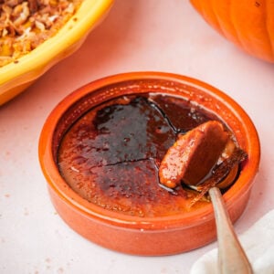 A ceramic ramekin filled with vegan pumpkin crème brûlée, showing its caramelized sugar topping being cracked with a spoon. The custard inside is creamy with a golden-brown top. A pumpkin and a pie are partially visible nearby.