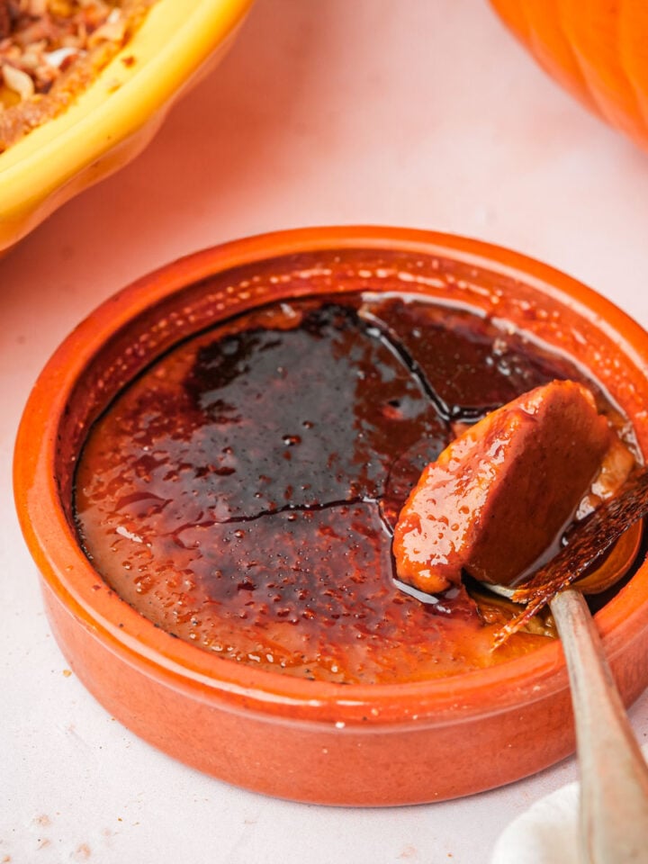 A ceramic ramekin filled with vegan pumpkin crème brûlée, showing its caramelized sugar topping being cracked with a spoon. The custard inside is creamy with a golden-brown top. A pumpkin and a pie are partially visible nearby.