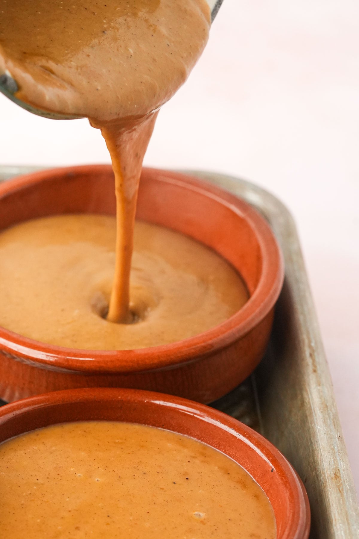 Vegan pumpkin creme brulee custard is being poured from a bowl into a round brown ramekin, which is placed on a metal baking sheet next to another filled ramekin.