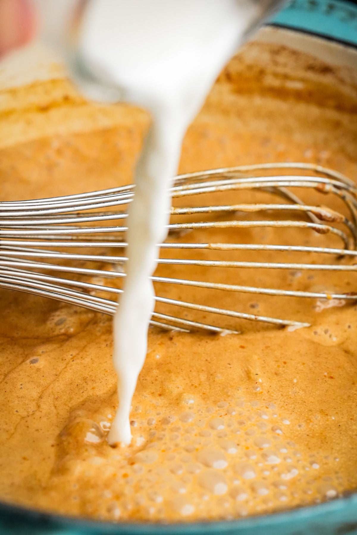 A close-up of starch slurry being poured into a pot of vegan pumpkin creme brulee custard with a metal whisk, as the mixture bubbles and blends together.