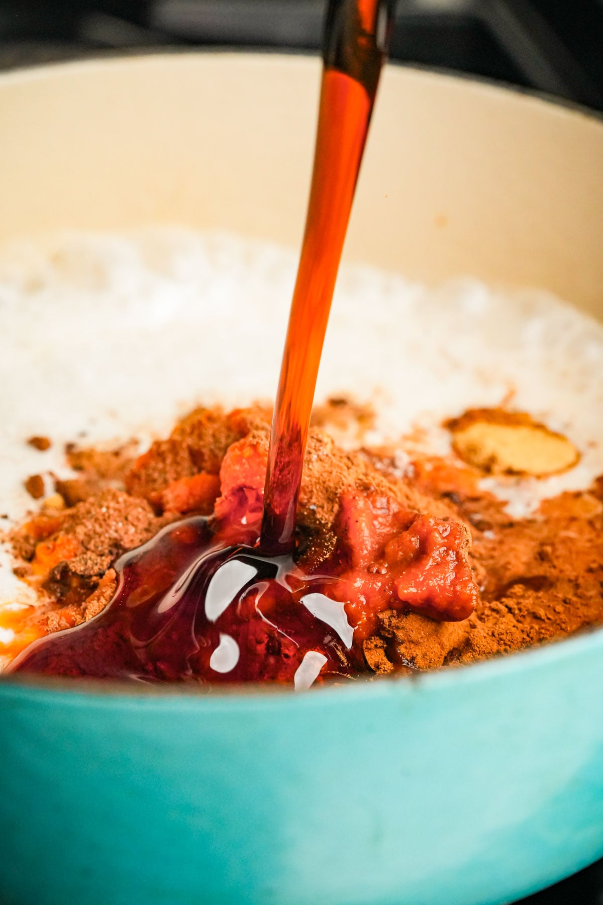 A close-up of a pot with frothy coconut milk, pumpkin puree spices, and a stream of dark maple syrup being poured in.