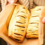 Two golden Gregg's-style vegan sausage rolls with visible meat filling are held in brown parchment paper. The pastries have slits on top, showing the filling inside, and are being held by a person in a beige, textured sweater.