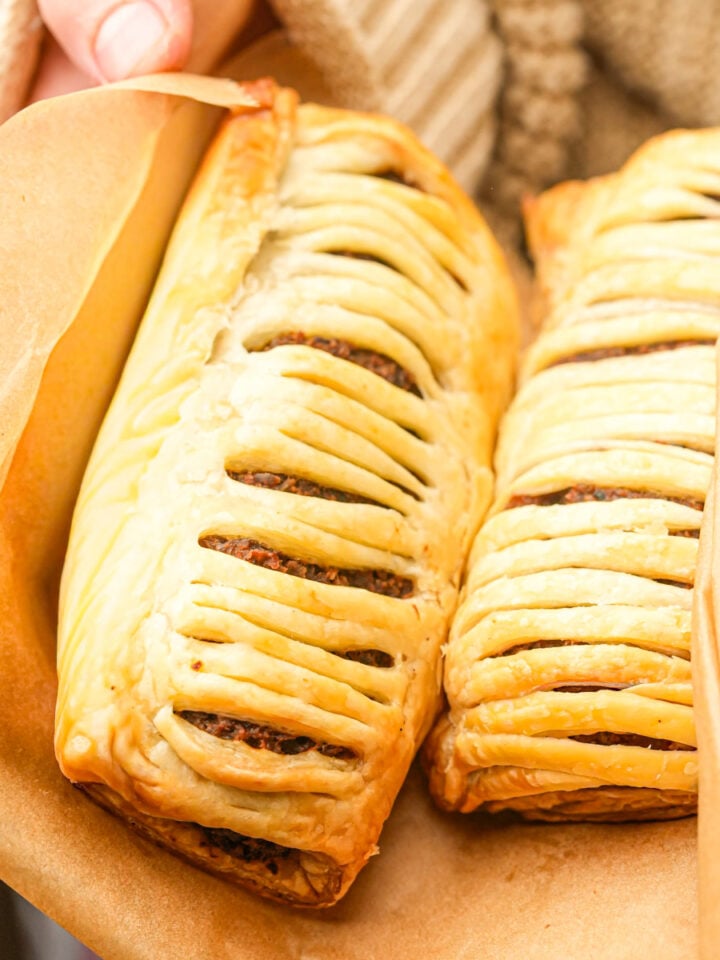 Two golden Gregg's-style vegan sausage rolls with visible meat filling are held in brown parchment paper. The pastries have slits on top, showing the filling inside, and are being held by a person in a beige, textured sweater.