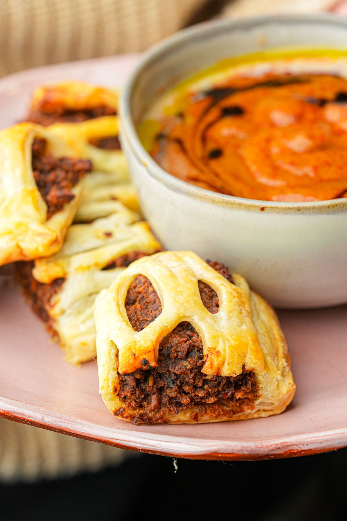 Golden vegan sausage rolls filled with a dark savory mixture are arranged on a pink plate next to a bowl of creamy orange soup garnished with oil and spices.