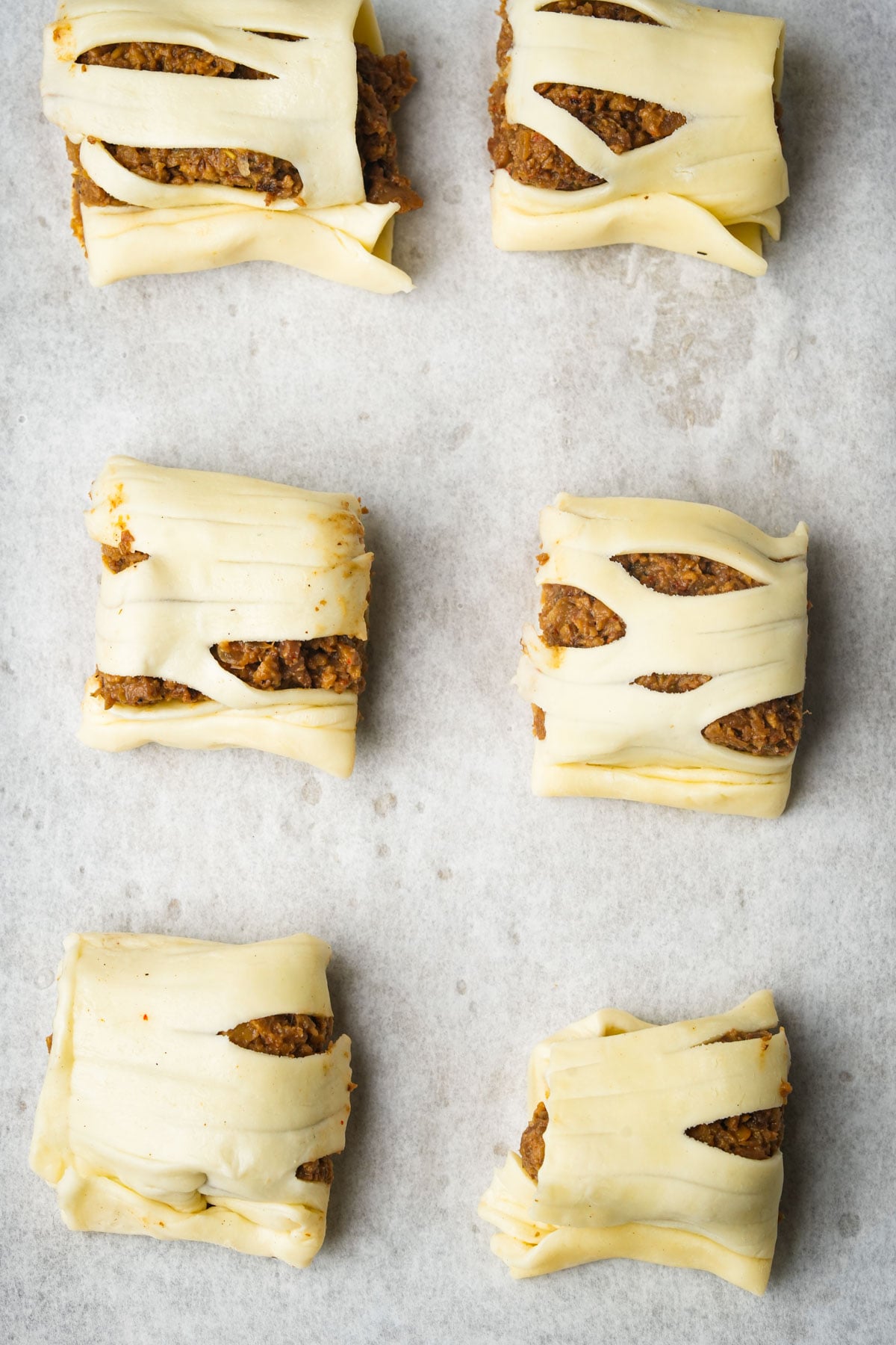 Six unbaked vegan sausage rolls filled with a spiced meat mixture are arranged in rows on a sheet of parchment paper. The pastry is sliced and folded to reveal the filling inside each parcel.
