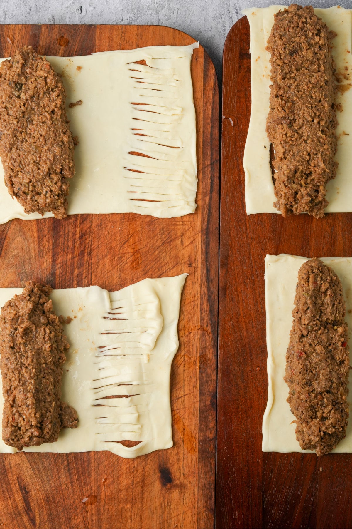 Four rectangles of puff pastry on wooden boards, each topped with a portion of brown mushroom filling; the pastry on the left has slits cut into it, ready to be wrapped around the filling to become vegan sausage rolls.