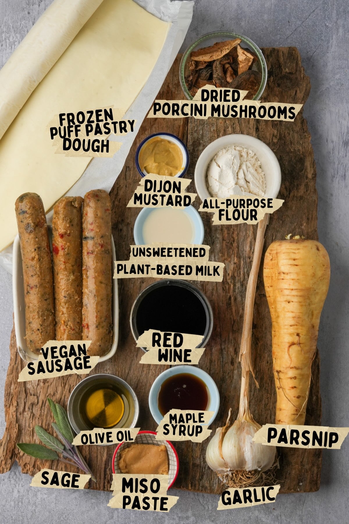 A wooden board displays ingredients for a vegan sausage rolls recipe: frozen puff pastry dough, dried porcini mushrooms, all-purpose flour, dijon mustard, vegan sausages, plant-based milk, red wine, olive oil, maple syrup, a parsnip, garlic, miso paste, and sage leaves.