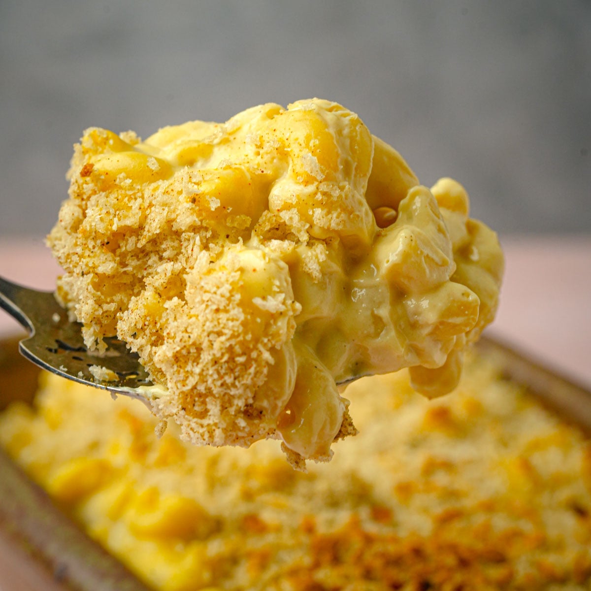 A close-up of a forkful of creamy vegan truffle mac and cheese, topped with golden, crispy breadcrumbs, held above a baking dish filled with more rich macaroni and cheese.