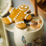A mug of vegan gingerbread latte topped with toasted marshmallows and a cinnamon stick, with a smiling gingerbread cookie resting on the rim. The mug has a floral design and sits on a table with a vintage plate nearby.