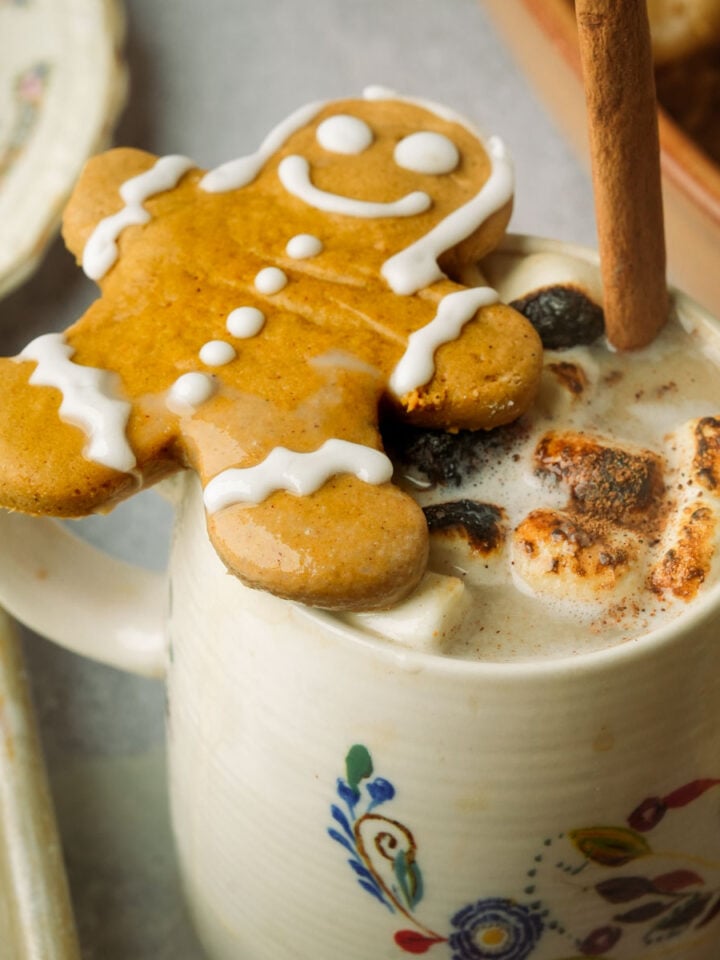 A mug of vegan gingerbread latte topped with toasted marshmallows and a cinnamon stick, with a smiling gingerbread cookie resting on the rim. The mug has a floral design and sits on a table with a vintage plate nearby.